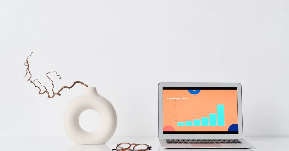 Un espace de travail minimaliste avec un ordinateur portable, un vase en céramique et des lunettes sur un bureau blanc.