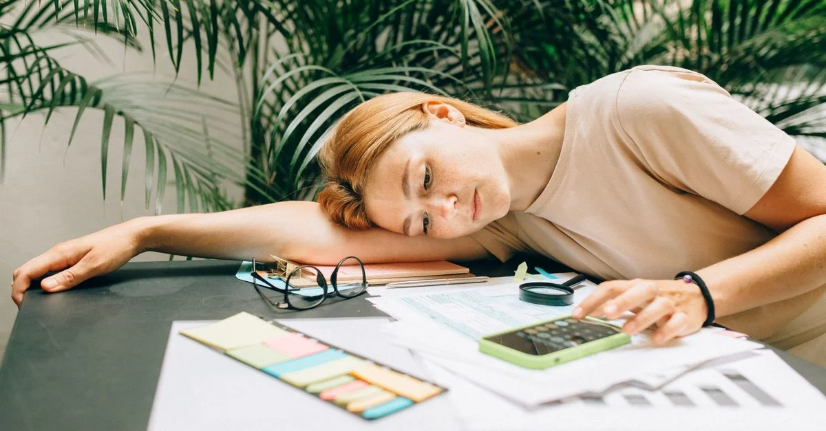 Une femme fatiguée calcule ses impôts à un bureau couvert de papiers et d'un smartphone.